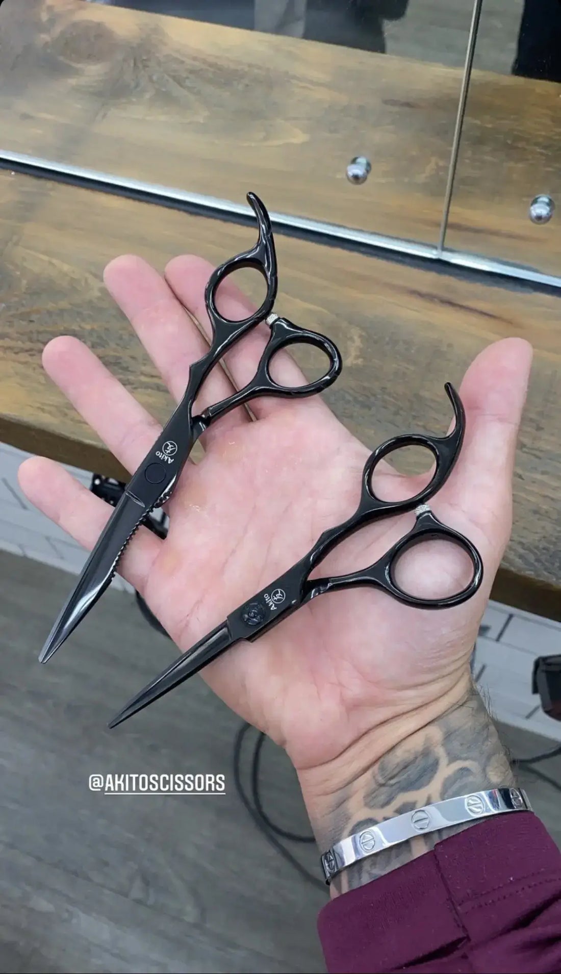 Hand holding two pairs of black akito scissors on a wooden surface