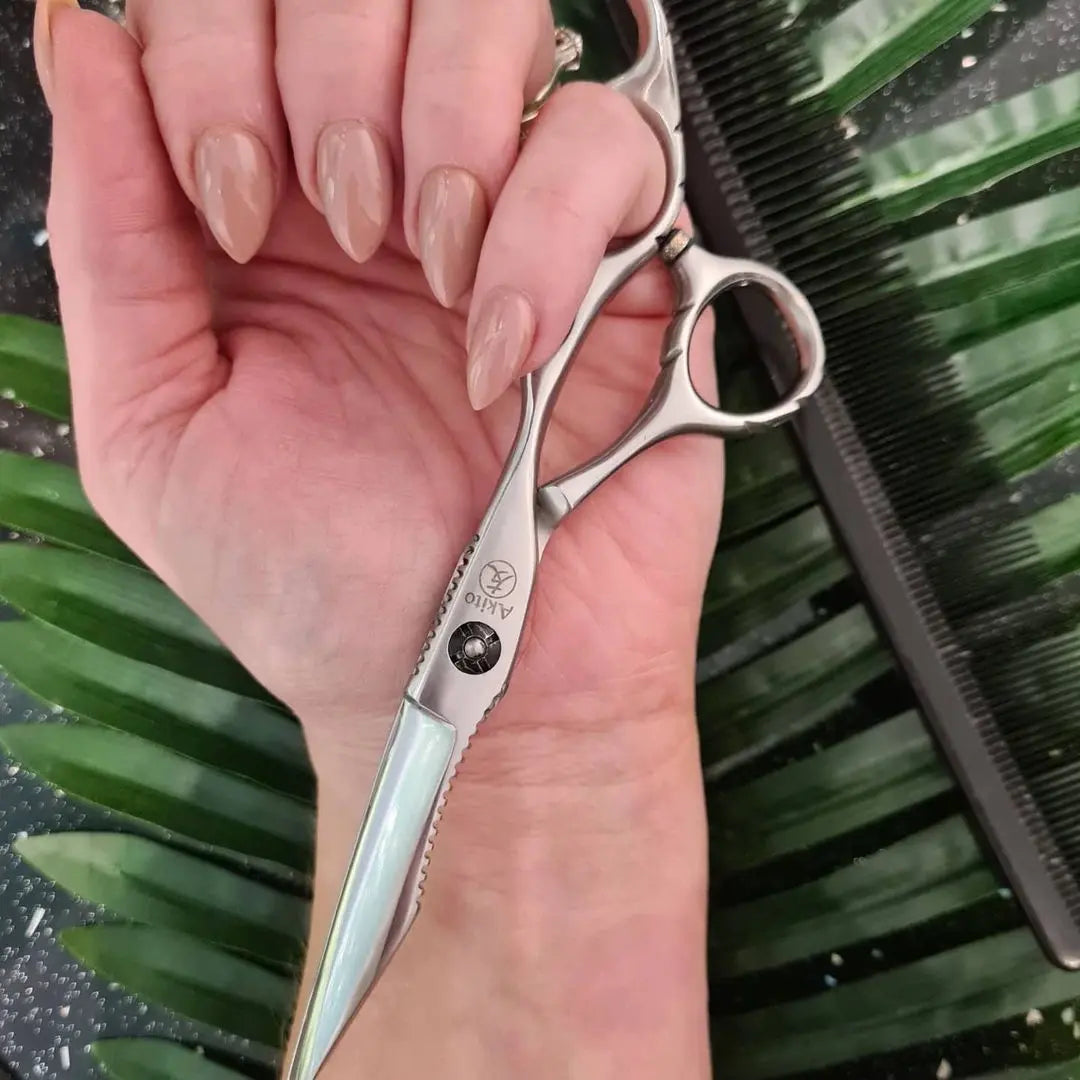 Hand holding a pair of silver akito scissors with a blurred green leaf background